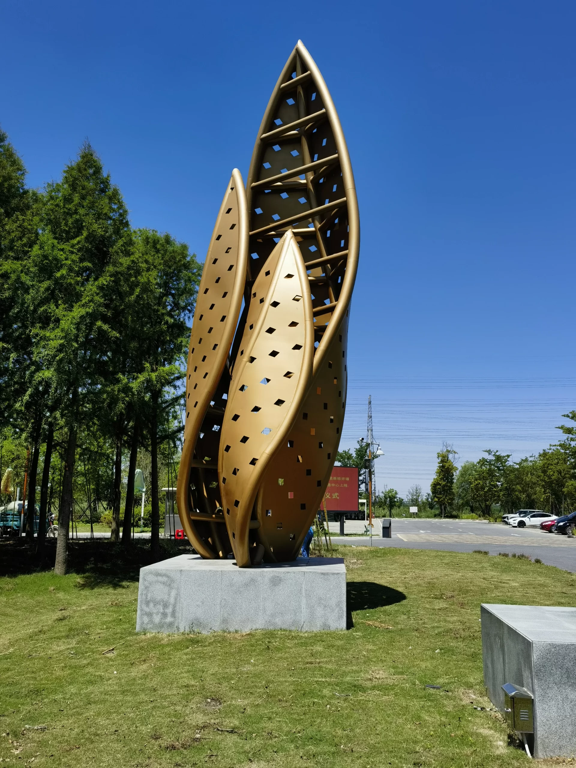Large gold leaf-shaped metal sculpture installed on a concrete base in a public park with landscape and partial person visible for scale