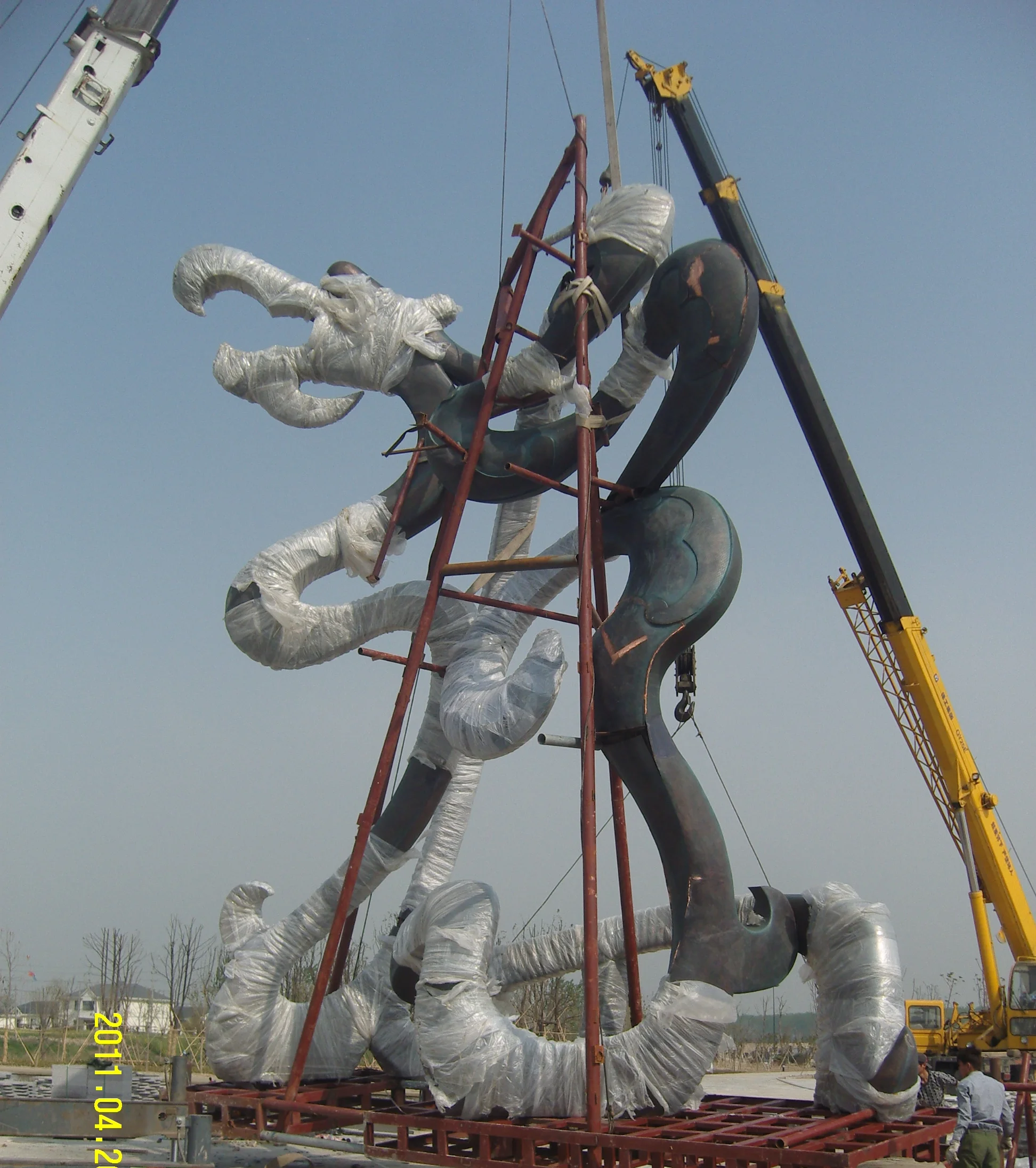 Workers using a crane to lift a large fiberglass dragon sculpture at an outdoor installation site, with workers visible for scale comparison