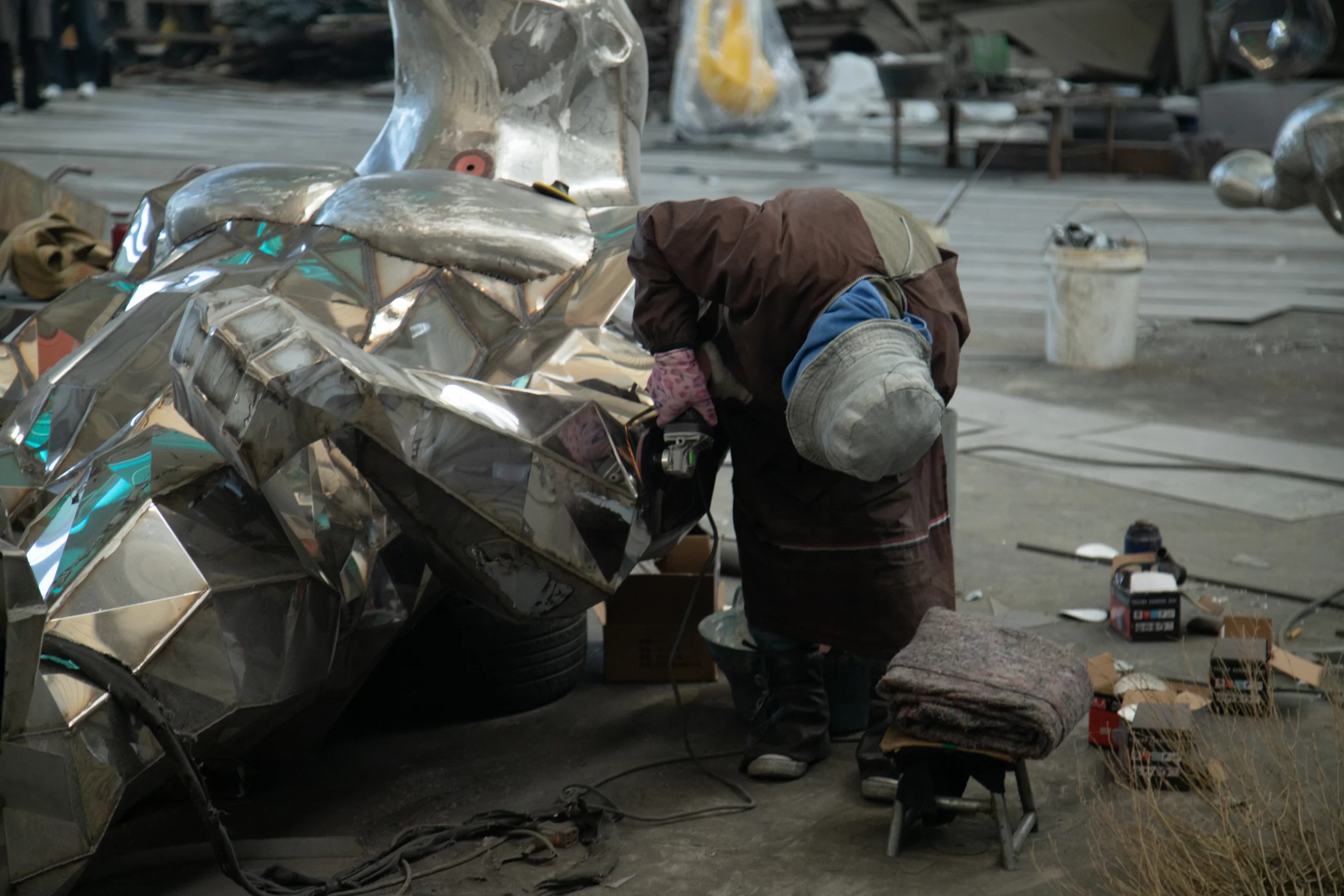 Factory worker in protective gear grinding and polishing a large faceted stainless steel sculpture in an industrial workshop
