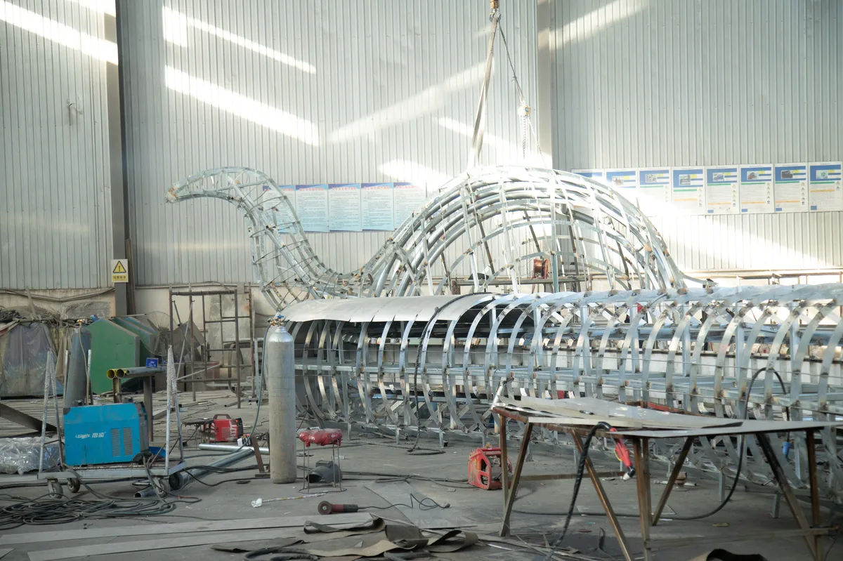 Spherical stainless steel sculpture assembly with overhead crane in manufacturing facility.